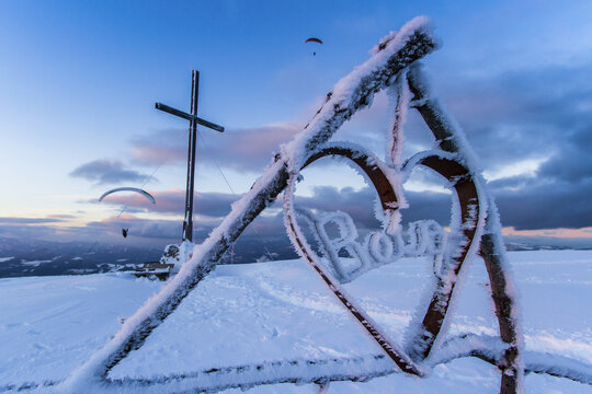 View of a snow-covered cross and heart-shaped structure with paragliders soaring in the distance, against a twilight sky, Schockl, Styria, Austria.