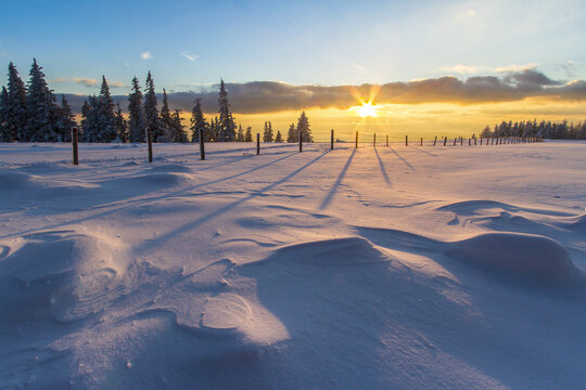 View of a snow-covered field with stark shadows cast by a rustic fence under a vibrant sunset sky, painting the landscape in warm and cool hues, Schockl, Styria, Austria.