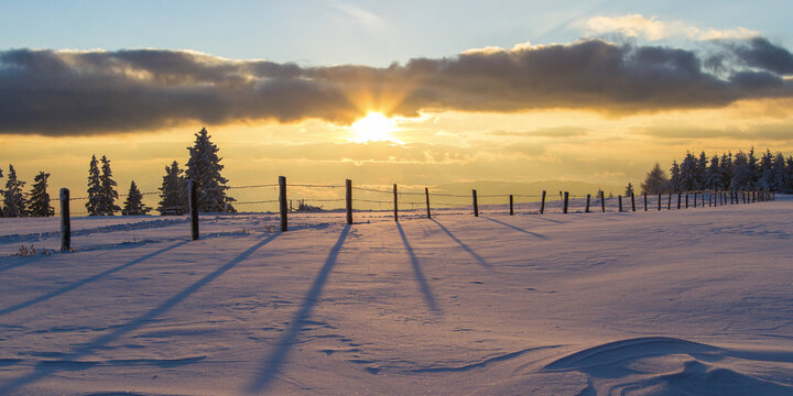 View of the sun bursts through clouds, casting long shadows from a fence across a snow-covered field lined with trees, Schockl, Austria.
