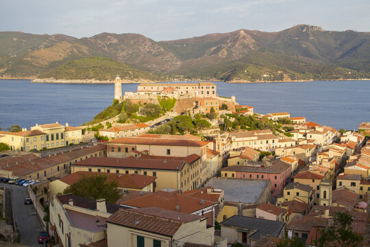 View of sun-kissed buildings with red-tiled roofs cascade down the hillside towards the shimmering sea, crowned by a historic fortress, Portoferraio, Elba, Tuscany, Italy.