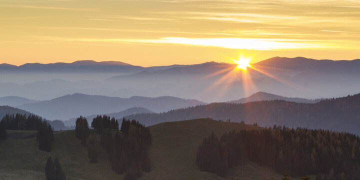 View of golden sunlight piercing through the hazy blue mountains, casting long shadows across the green hillsides, Sommeralm, Steiermark, Austria.