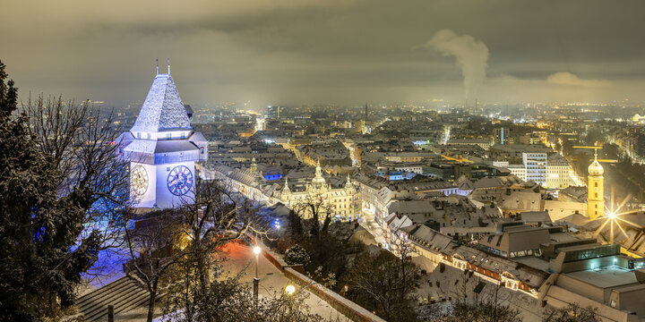 View of illuminated clock tower standing proudly amidst a snow-dusted cityscape under a night sky, its face glowing warmly, Graz, Styria, Austria.