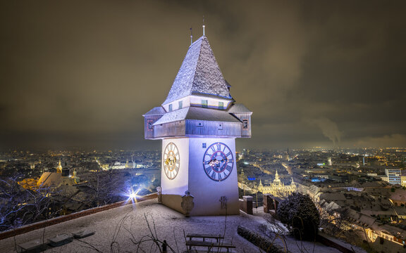View of the iconic clock tower, the Uhrturm, standing majestically against the twilight sky, casting a warm glow over the snow-dusted city, Graz, Styria, Austria.