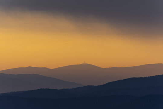 View of layered mountain silhouettes fading into the golden sky, a distant peak crowned with a structure, casting long shadows in Brandlalm, Styria, Austria.