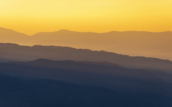 View of layered mountain ranges fading into the horizon under a warm, golden sky, creating a serene and majestic atmosphere, Brandlalm, Styria, Austria.