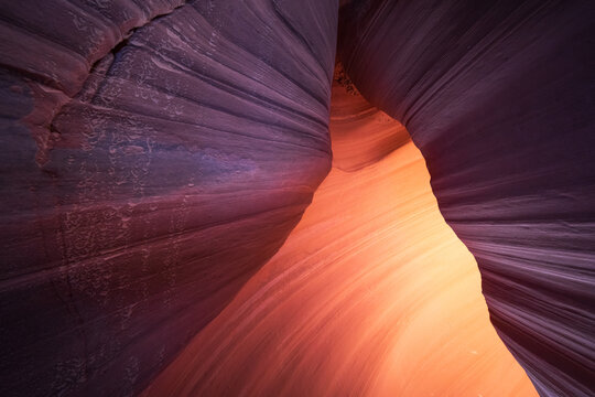 View of smooth, sculpted sandstone walls casting shadows, leading to a bright orange glow within the canyon's depths, Escalante, Utah, United States.