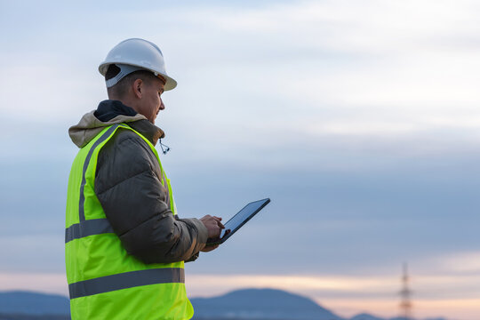 A young male electrical engineer works outdoors, inspecting high-voltage power lines with a tablet in a utility area at dusk.