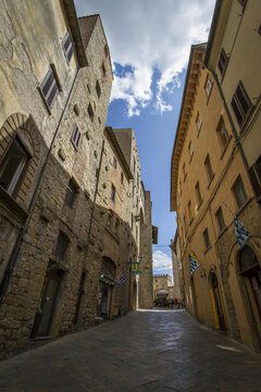 View of aged stone buildings cast long shadows on a narrow cobblestone street under a radiant blue sky, Volterra, Tuscany, Italy.