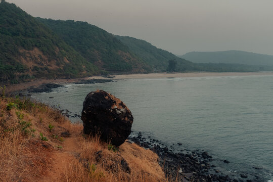 View of a serene coastal landscape with a massive dark boulder perched on a grassy cliff edge overlooking the tranquil waters, Chiplun, Maharashtra, India.