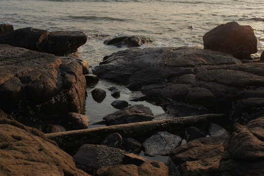 View of sun-kissed rocks meet the gentle waves in a serene coastal landscape, the tranquil water reflecting the sky, Chiplun, Maharashtra, India.