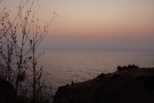 View of gentle waves lap against the shore under a soft, pastel sky, with silhouettes of dry plants and rocky cliffs framing the tranquil scene, Chiplun, Maharashtra, India.