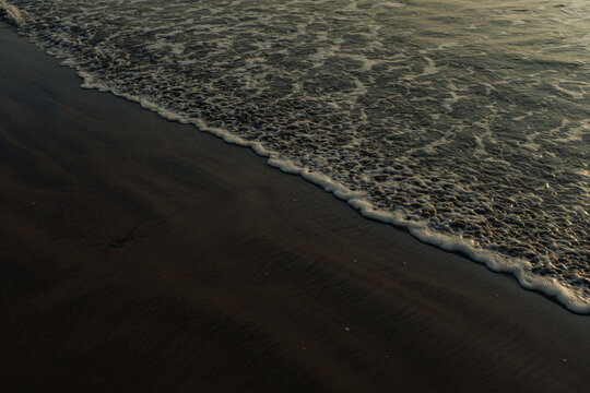View of dark sands meeting the foamy, frothy embrace of the ocean waves under the fading light, creating a serene coastal scene, Chiplun, Maharashtra, India.