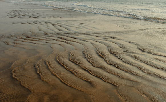 View of the wet sand with wavy patterns reflecting sunlight as the tide comes in, creating a serene coastal scene, Chiplun, Maharashtra, India.