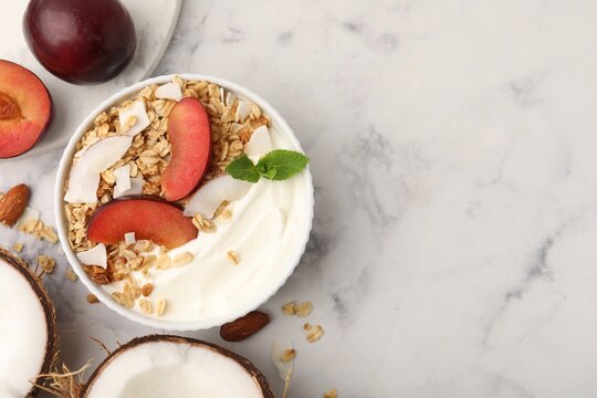 Delicious coconut yogurt with granola and plum in bowl on light marble table, flat lay. Space for text