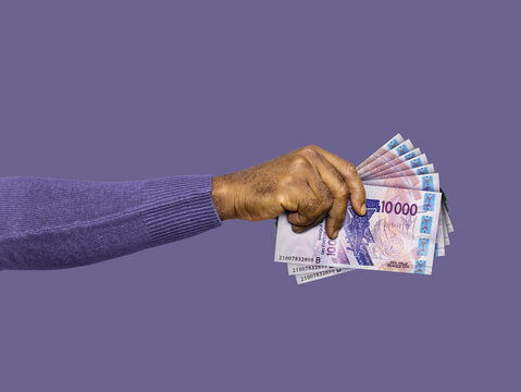 A hand in a purple sweater holds a fan of 10,000 West African CFA franc notes against a purple background. A clean, conceptual shot representing African finance, wealth, and currency exchange.