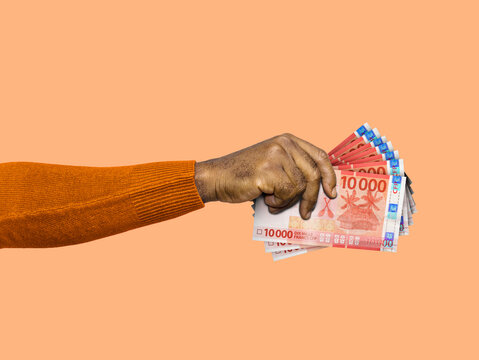 A hand in an orange sweater holds a fan of red 10,000 CFP franc notes. The image features Polynesian currency and cultural icons against a clean, vibrant orange backdrop.
