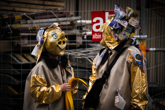 Basel, Switzerland - 10 March 2025: View of two masked figures, one with a pig-like visage, adorned in gold, standing against a backdrop of urban scaffolding and the 'albin' sign.
