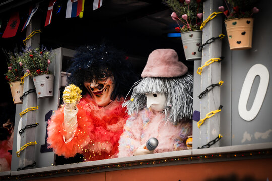 Basel, Switzerland - 10 March 2025: View of two masked figures in vibrant red and soft pink costumes, peering out from a decorated float, adorned with flowers and flags.