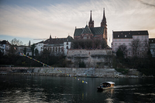 Basel, Switzerland - 10 March 2025: View of the majestic Basel Minster rising above the Rhine, its spires piercing the dusk sky, with a boat gliding along the water's surface.