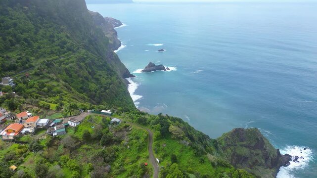 Aerial view of a vibrant Arco de Sao Jorge village nestled on the lush green cliffs of Madeira, with the Atlantic Ocean stretching to the horizon under a cloudy sky.