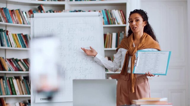 A young woman with light skin is teaching a math lesson. She stands in front of a whiteboard with equations, engaging with students in a classroom filled with bookshelves.