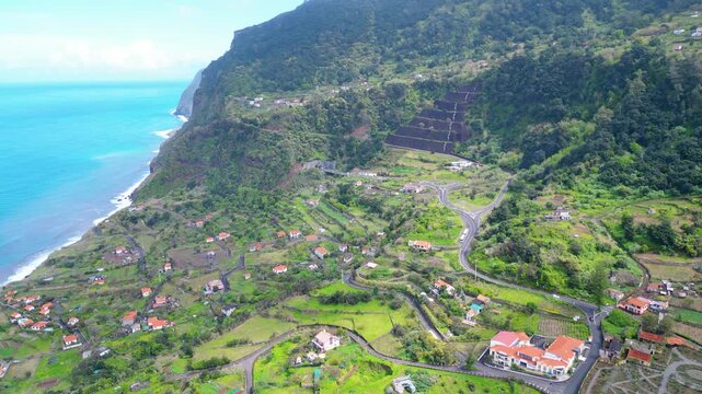 Aerial view of a vibrant Arco de Sao Jorge village nestled on the lush green cliffs of Madeira, with the Atlantic Ocean stretching to the horizon under a cloudy sky.