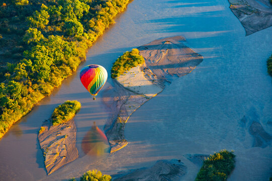 Albuquerque, United States - 11 October 2017: View of a hot air balloon drifting over the Rio Grande, its vibrant colors mirroring the golden hues of the autumnal trees dotting the riverbank.
