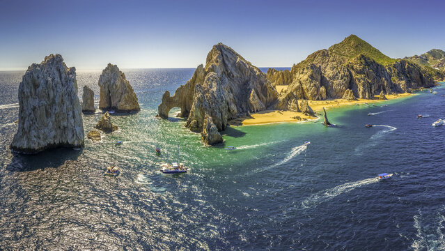 Aerial view of El Arco's striking rock formations meet the turquoise sea, boats gliding, and golden sands shimmering under the sun, Cabo San Lucas, Mexico.