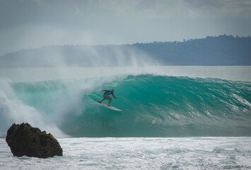 Obraz premium Male surfer rides a fast and hollow turquoise wave with offshore mist. Exposed sharp rock in the foreground shows danger of surfing reef breaks in tropical paradise of Mentawai Islands in Indonesia.