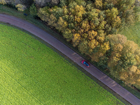 Aerial view of a lone red car traversing the road, flanked by a vibrant green field and a forest with autumn hues, Diever, Drenthe, Netherlands.
