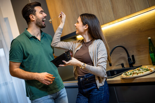 Happy young couple playing and laughing while eating pasta in the kitchen.