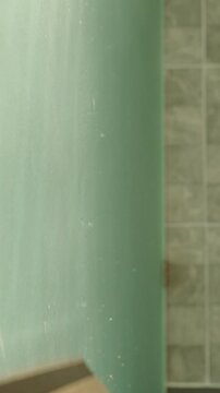 Vertical video. Person Using Squeegee on Steamy Glass Surface to Remove Moisture in Bright Bathroom