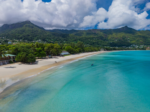 Aerial view of the pristine white beach meeting the turquoise waters, backed by lush green mountains under a partly cloudy sky, Victoria, Seychelles.