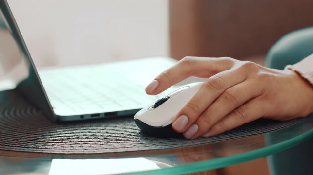A woman uses a mouse while working on a laptop. The setting is well lit and modern, featuring a glass table. The focus is on productivity and technology use in everyday life.