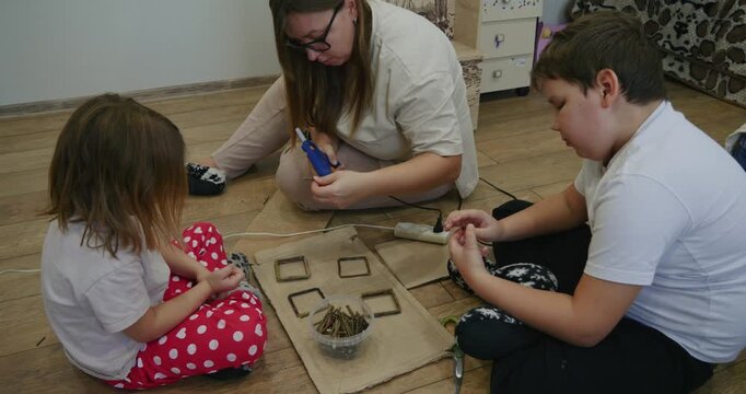 Happy family engaging in a creative project, making handmade picture frames from small wooden sticks and a hot glue gun while sitting together on the floor, fostering teamwork and creativity.