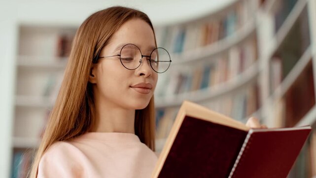 A young woman with long brown hair reads a book in a library. She wears glasses and focuses on the text. Bookshelves filled with books are seen in the background.