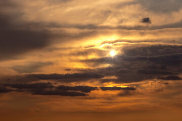 Zachmurzone, baśniowe niebo o zachodzie słońca z warstwami ciemnych i jasnych chmur tworzy dramatyczny efekt. Słońce jest częściowo przesłonięte przez chmury, rzucając ciepłe, złote światło na scenę.  © Grzegorz