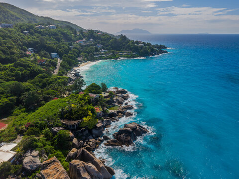 Aerial view of turquoise waters meeting the lush green coastline near granite boulders, with buildings nestled among the trees, La Retraite, Mahe Island, Seychelles.