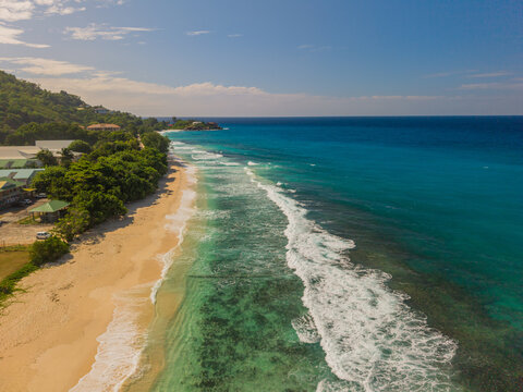 Aerial view of turquoise waters gently kiss the sandy shore, framed by verdant trees against a clear blue sky, La Retraite, Mahe Island, Seychelles.