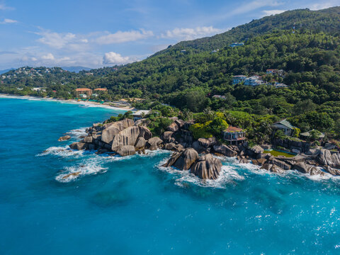 Aerial view of turquoise waters meeting the rocky coastline, lush hills dotted with homes, creating a vibrant contrast of blues and greens, La Retraite, Mahe Island, Seychelles.