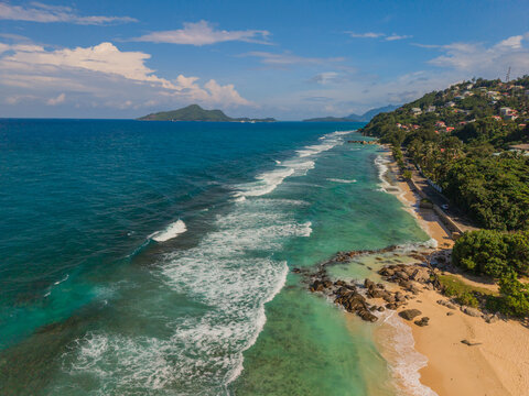 Aerial view of turquoise waves gently kissing the golden sands, lush green hills meeting the azure sky, La Retraite, Mahe Island, Seychelles.