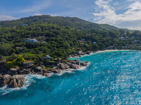Aerial view of azure waves crash against the rocky shore beneath lush green hillsides, La Retraite, Mahe Island, Seychelles.