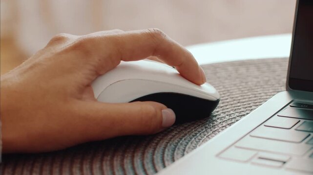 A hand operates a white computer mouse placed on a textured mat. The scene shows a laptop nearby, indicating an active work or study session in a cozy setting.