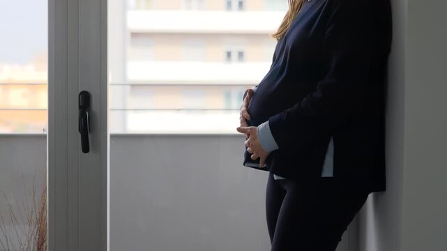 Side profile of pregnant woman in dark blazer standing beside a large window, hands cradling bump, city building visible outside, natural light