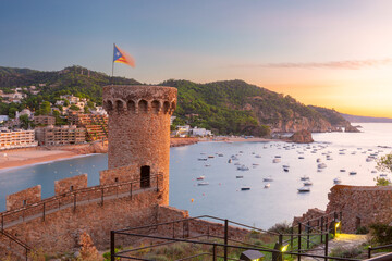 Historic stone watchtower overlooking the calm bay of Tossa de Mar, Costa Brava, Spain, with anchored boats and warm sunset light over the Mediterranean coast.