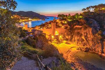 Elevated night view of the fortified old town of Tossa de Mar, Spain, with illuminated stone walls, towers, and the calm Mediterranean bay under deep blue twilight.