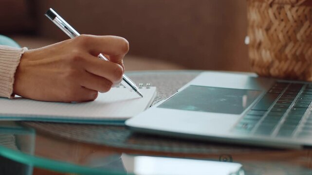 A woman with light skin writes notes in a notebook using a pen. A laptop is open on the table in front of her. The setting is indoors with natural light.
