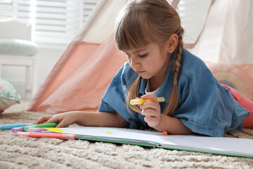 Little girl drawing in sketchbook near toy wigwam at home © New Africa