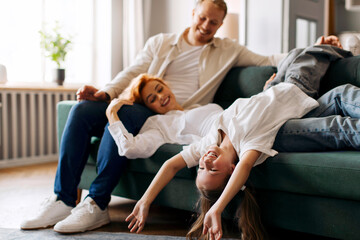 Excited family of three relaxing on comfortable couch, sharing joyful moments, girl playfully...