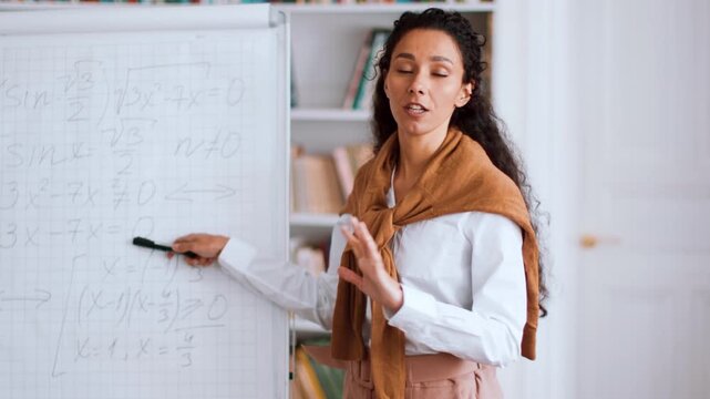 A woman with medium skin tone and long curly hair stands in front of a whiteboard, teaching math concepts. She holds a marker and engages with students in a bright classroom.